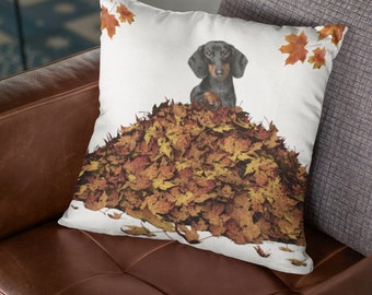 May include: A white throw pillow with a black and brown dachshund sitting in a pile of autumn leaves. The leaves are orange, yellow, and brown. There are also a few red leaves on the pillow.