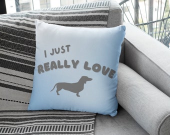Puede incluir: Una almohada de color azul claro con el texto "I just really love" en gris, con una silueta de un dachshund en gris. La almohada está sobre un sofá de rayas grises y blancas.