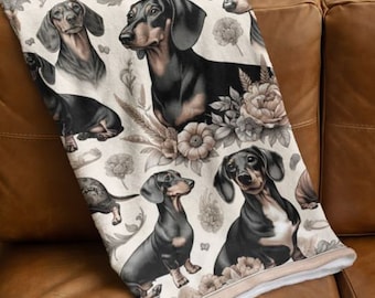 May include: A white throw blanket with a repeating pattern of black dachshund dogs and floral designs. The dogs are depicted in various poses, including sitting, standing, and running. The floral designs are in shades of pink, purple, and green.