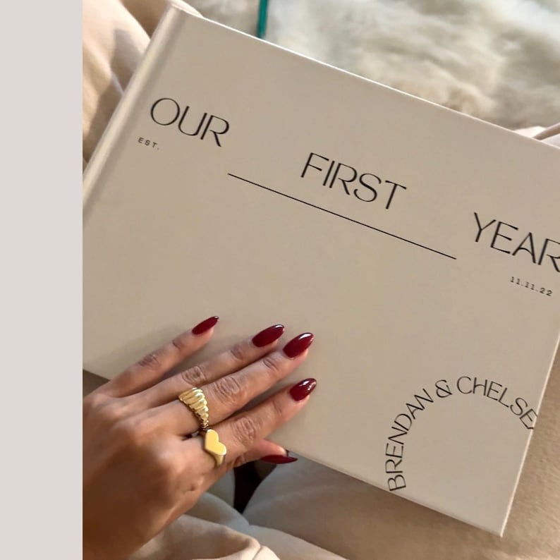 May include: A white hardcover book with the text "OUR FIRST YEAR 11.11.22 BRENDAN & CHELSEY" printed on the cover.