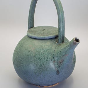 Puede incluir: Tetera de cerámica hecha a mano con cuerpo redondeado y asa curva. Esmaltada en tonos azules y verdes, con superficie texturizada. La tapa tiene un pequeño asa y un pico para verter.
