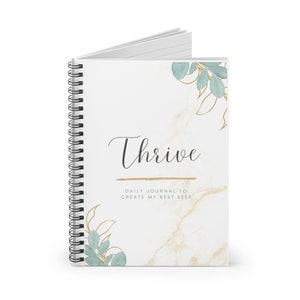 Puede incluir: Un cuaderno blanco de espiral con un patrón de mármol y detalles dorados. La portada presenta la palabra "Thrive" en letra cursiva negra y el texto "Diario para crear mi mejor yo". El cuaderno está decorado con hojas verdes de acuarela.