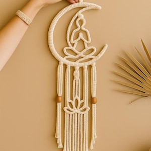 Op de afbeelding: Crèmekleurige macramé wandhanger met een halve maan ontwerp en een zittende figuur in het midden. Het stuk bevat een lotusbloem en houten kralen, met geknoopte franjes aan de onderkant. Een hand houdt de bovenkant van de wandhanger vast.