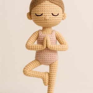 May include: A handmade crochet doll in a yoga pose. The doll has a light brown bun, closed eyes, and is wearing a pink leotard. The doll is standing on one leg with hands together in a prayer position. The doll is made of beige and pink yarn.