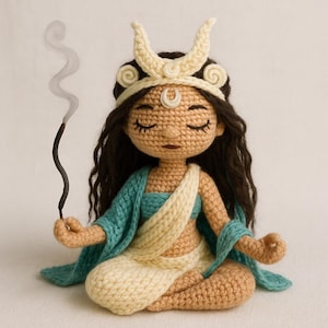 May include: Crocheted doll in a meditative pose, holding an incense stick with smoke. The doll has long dark hair, closed eyes, and wears a cream-colored crescent moon crown, a teal shawl, and cream-colored and tan clothing. The doll is sitting cross-legged.
