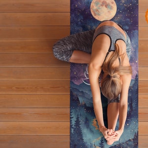 May include: A woman practices yoga on a blue and purple yoga mat with a moon and forest scene. The mat has a gold foil accent and the text "Goldenlia" printed on it.