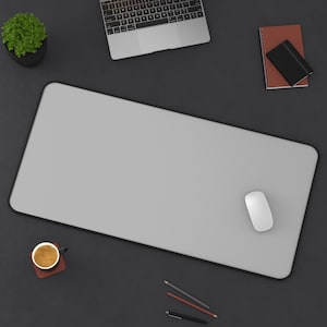 Pu&ograve; includere: Vista dall'alto di un'area di lavoro con un tappetino per mouse grigio con bordo nero, un mouse bianco, un laptop, una piccola pianta in vaso, un taccuino, una tazza di caff&egrave; e matite. Su una superficie grigio scuro.