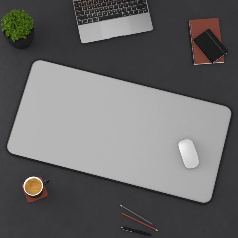 Pu&ograve; includere: Vista dall'alto di un'area di lavoro con un tappetino per mouse grigio con bordo nero, un mouse bianco, un laptop, una piccola pianta in vaso, un taccuino, una tazza di caff&egrave; e matite. Su una superficie grigio scuro.