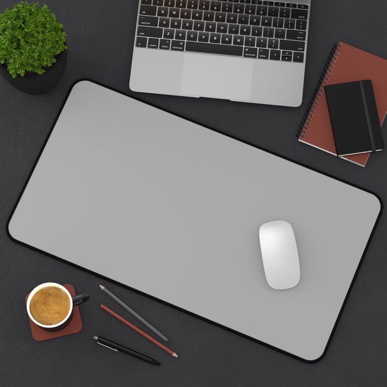 Pu&ograve; includere: Vista dall'alto di uno spazio di lavoro con un tappetino per mouse grigio con bordo nero, un mouse bianco, un laptop, una tazza di caff&egrave; e articoli di cancelleria. Sono presenti anche una piccola pianta in vaso e quaderni.