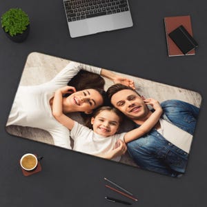 Pu&ograve; includere: Tappetino per mouse rettangolare con un ritratto di famiglia. L'immagine mostra una famiglia sorridente di tre persone sdraiate su un pavimento di legno. Il tappetino per mouse &egrave; circondato da una superficie nera, un laptop, un taccuino, una tazza di caff&egrave; e matite.