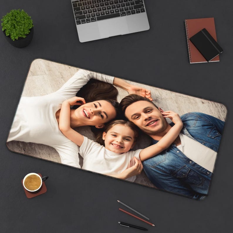Pu&ograve; includere: Tappetino per mouse rettangolare con un ritratto di famiglia. L'immagine mostra una famiglia sorridente di tre persone sdraiate su un pavimento di legno. Il tappetino per mouse &egrave; circondato da una superficie nera, un laptop, un taccuino, una tazza di caff&egrave; e matite.