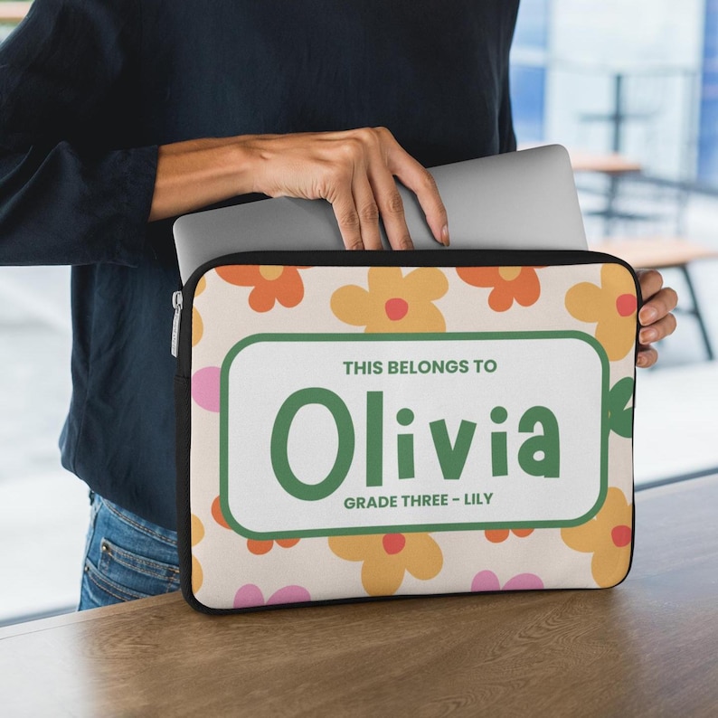 May include: A beige laptop sleeve with a floral pattern in orange, yellow, and pink. The sleeve has a green and white rectangular label that reads "THIS BELONGS TO Olivia GRADE THREE - LILY".