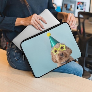 May include: A laptop sleeve featuring a light blue background with a brown dog wearing yellow star-shaped glasses and a green party hat. The sleeve has a black border and is held with a laptop.