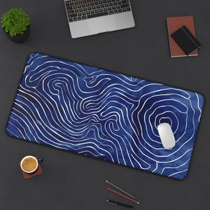 May include: A blue and white desk mat with a topographic design. The mat is rectangular and features a pattern of white lines on a blue background. A white computer mouse rests on the mat, along with a laptop, coffee cup, and other office supplies.