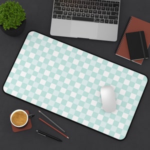 May include: A green and white checkered mouse pad with a white computer mouse on top. The mouse pad is on a black surface with a laptop, a coffee cup, pencils, and a notebook.