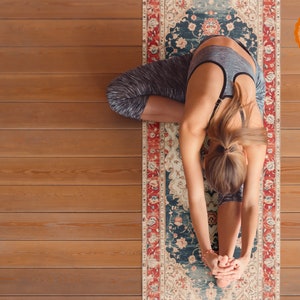 May include: A woman in a grey sports bra and black leggings is doing a yoga pose on a patterned yoga mat. The mat has a red, blue, and white design with floral patterns. The mat has a logo in the corner that says "GOLDELIA MEDITATION & YOGA".