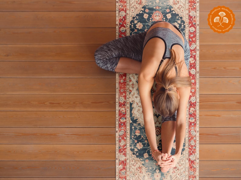 May include: A woman in a grey sports bra and black leggings is doing a yoga pose on a patterned yoga mat. The mat has a red, blue, and white design with floral patterns. The mat has a logo in the corner that says "GOLDELIA MEDITATION & YOGA".