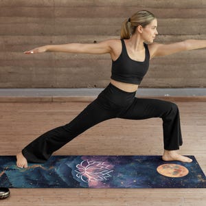 May include: A woman in black yoga pants and a black tank top is doing a yoga pose on a dark blue yoga mat with a galaxy print. The mat has a pink and white lotus flower design and a large orange moon design. Two small metal singing bowls are on the floor next to the mat.