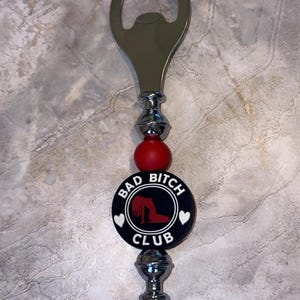 Könnte beinhalten: Ein silberner Flaschenöffner mit einem roten und schwarzen Perlen-Griff. Die Perle hat einen schwarzen Kreis mit weißem Text, der "Bad Bitch Club" lautet, und eine rote Silhouette einer Frau in einem Kleid.