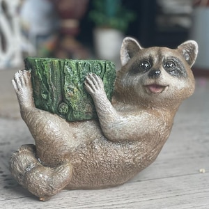 May include: A whimsical raccoon figurine holding a green tree stump planter. The raccoon has brown and tan fur with a black mask around its eyes. The planter is textured with a wood grain design.
