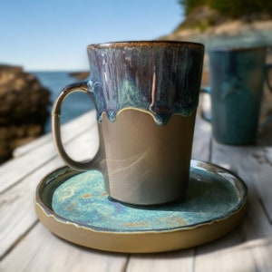 May include: A brown ceramic coffee mug with a teal glaze and a drip effect. The mug is sitting on a matching teal saucer.