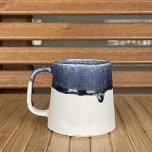 Könnte beinhalten: Eine weiße Keramiktasse mit blau-weißem Glase. Die blaue Glasur hat einen gesprenkelten Effekt und tropft an der Seite der Tasse herunter.