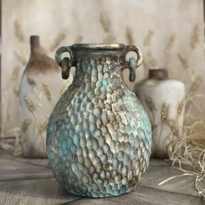 Op de afbeelding: Een decoratieve keramische vaas met een textuur, gehamerde afwerking in tinten teal, bruin en goud. De vaas heeft twee handvatten en een rond lichaam.