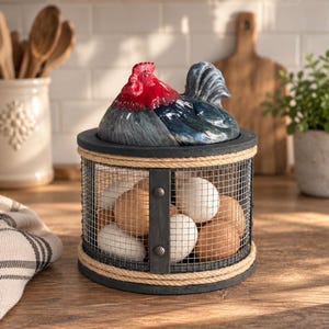 May include: A cylindrical egg basket with a wire mesh body, black metal frame, and natural rope trim. The lid is a ceramic cockerel figurine with a bright red comb and dark blue feathers. The basket is filled with several white and brown eggs.