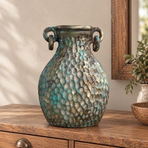 May include: A bulbous vase with a textured, hammered surface featuring a weathered finish in shades of metallic bronze and oxidized teal. The vessel has a wide neck and two small, circular handles attached to the upper rim on opposite sides.