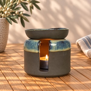 May include: A cylindrical ceramic oil burner with a matte dark gray base and a glossy blue and yellow reactive glaze rim. The burner features a wide central opening for a tea light candle and a shallow dish top for wax melts or essential oils.