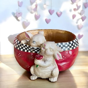 May include: A red and white checked planter with two white ceramic bunnies peeking out. The bunny on the right is holding a red flower.