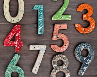 May include: A collection of ceramic house numbers, each with a unique color and texture. The numbers range from zero to nine, with colors including green, red, orange, and blue. Each number is approximately 6 inches tall.