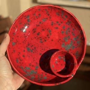 Op de afbeelding: Rode keramische serveerschaal met een kleinere dipschaal eraan bevestigd. De schaal heeft een gespikkeld patroon met zwarte en grijze vlekken.