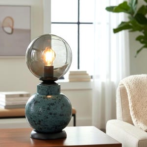 Könnte beinhalten: Dekorative Tischlampe mit tealfarbenem, gesprenkeltem Sockel und einem transparenten, kugelförmigen Schirm. Die Lampe hat eine warme, leuchtende Glühbirne und einen schwarzen Sockel. Die Lampe ist ca. 30 cm hoch.
