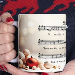 May include: White ceramic mug with a Christmas-themed design. The mug features a close-up of a red and gold ornament with a background of sheet music for the song "Silent Night".