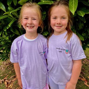 May include: Two young girls wearing matching lavender t-shirts and shorts. The shirts have the text "Werth y byd i gyd" and a small graphic. One girl wears a pink and white striped skirt, the other wears lavender shorts. They are smiling in front of green foliage.