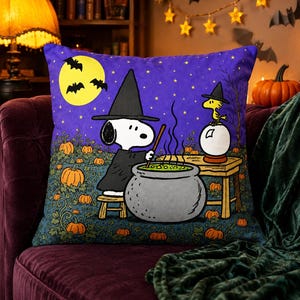 May include: A square throw pillow featuring a purple night sky with a yellow moon and bats. Snoopy wears a black witch hat and stirs a green bubbling cauldron. Woodstock sits on a crystal ball on a wooden table in a pumpkin patch.