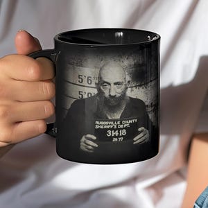 May include: Black ceramic mug featuring a black and white image of a man's mugshot. The mug displays the man's height, and the text "RIDGEVILLE COUNTY SHERIFF'S DEPT" and "31418 26 77".