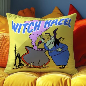 May include: A yellow decorative pillow with a cartoon witch illustration. The witch is stirring a cauldron with the text "WITCH HAZEL" above. A black cat is to the left of the cauldron. The pillow is on a yellow couch with orange and red pillows.