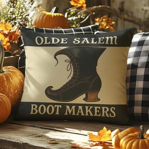 May include: A decorative pillow with the text "OLDE SALEM BOOT MAKERS" and a black boot illustration. The pillow is black with a cream-colored center. Pumpkins and fall foliage are in the background.