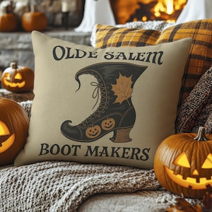 May include: Beige throw pillow with the text "Olde Salem Boot Makers" and a graphic of a black witch's boot with a maple leaf and two jack-o'-lanterns. The pillow is surrounded by pumpkins and fall-themed decor.