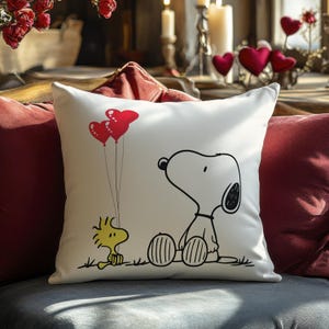 May include: White decorative pillow featuring Snoopy and Woodstock. Snoopy is sitting, and Woodstock is holding two red heart-shaped balloons. The pillow is on a dark gray surface, with red pillows in the background.