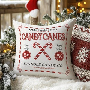 May include: White square pillow with a red and white candy cane design. The pillow features the text "Olde Fashioned Candy Canes" and "Kringle Candy Co." with crossed candy canes and the price "5¢".