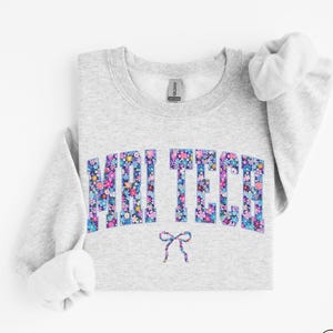 May include: Ash-colored sweatshirt with the text "MAI TECH" in a floral pattern. A small floral bow is below the text. The sweatshirt has a crew neck and long sleeves.