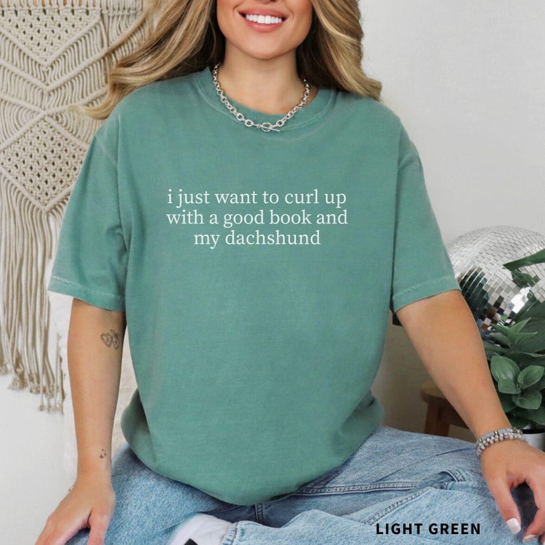 Puede incluir: Camiseta verde claro con el texto blanco: "i just want to curl up with a good book and my dachshund". La camiseta tiene cuello redondo y mangas cortas. Las palabras "LIGHT GREEN" est&aacute;n en la parte inferior de la imagen.