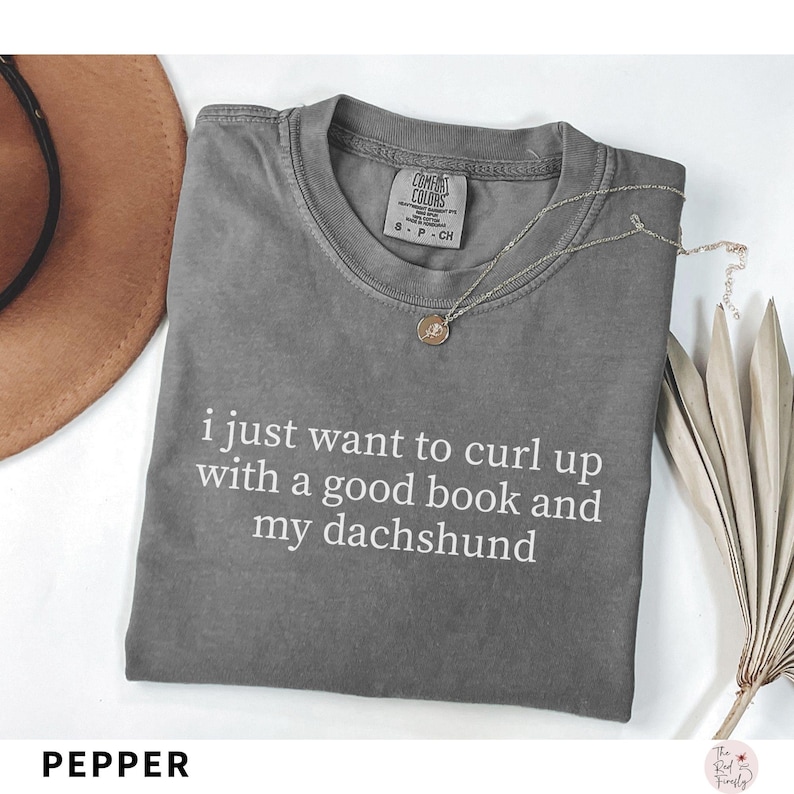 Puede incluir: Camiseta gris con el texto blanco "i just want to curl up with a good book and my dachshund". Un collar plateado con un colgante descansa sobre la camiseta. Un sombrero marr&oacute;n y una planta seca est&aacute;n en el fondo.