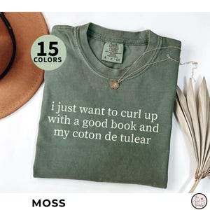 May include: A moss green t-shirt with the text "i just want to curl up with a good book and my coton de tulear" in white. A gold necklace is draped on the shirt. The shirt is from Comfort Colors.