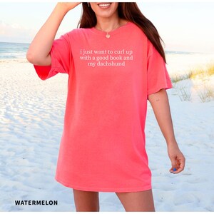 Puede incluir: Una camiseta de color sand&iacute;a con el texto "i just want to curl up with a good book and my dachshund" en blanco. La camiseta es de corte holgado y parece estar hecha de un material suave. La palabra "WATERMELON" est&aacute; impresa en la parte inferior.