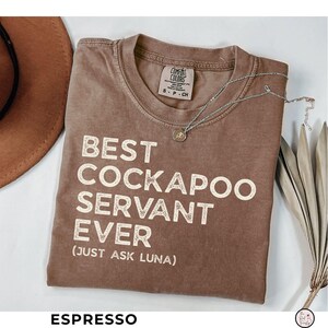 May include: A white ceramic mug with a black handle and the text "But First, Coffee" in a bold, black font. The mug is set against a blurred background, suggesting a cozy setting.