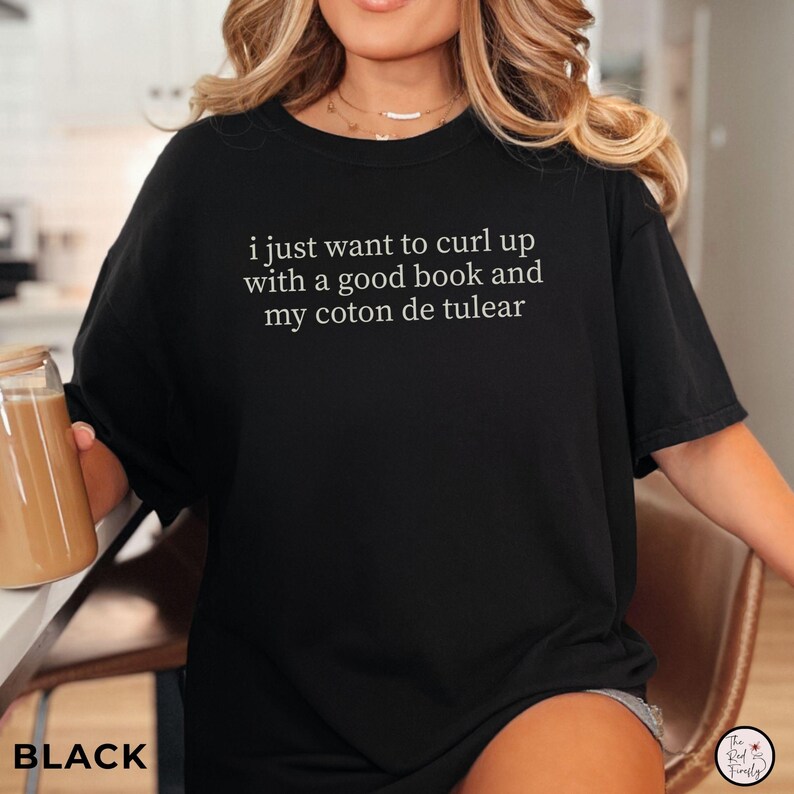 May include: Black t-shirt with white text: "i just want to curl up with a good book and my coton de tulear". The shirt has a loose fit and short sleeves. The word "BLACK" is printed at the bottom left of the image.